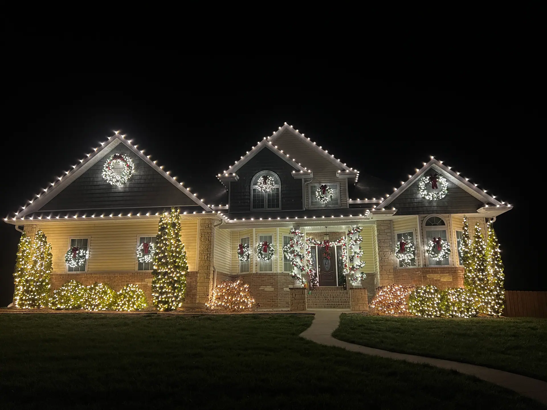 A home lit at night with warm white lights along the gable edges, lit wreaths in the windows and glowing shrubs across the front